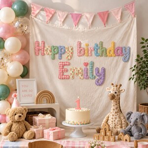 May include: A cream-colored fabric backdrop features the text Happy Birthday Emily in colorful, patterned, stitched letters. Below, a white cake with a number one candle sits on a stand, surrounded by plush toys, gift boxes, and wooden blocks.