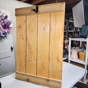 May include: A wooden wall-mounted coat rack with a natural finish. The rack features vertical wooden planks with a darker wood trim at the top and bottom. A black metal hook is attached to the top trim.