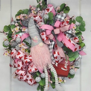 May include: A Valentine's Day wreath with a gnome figure. The wreath is decorated with pink and red heart-patterned ribbons, pink bows, and artificial foliage. A small red sign displays the word "Love".