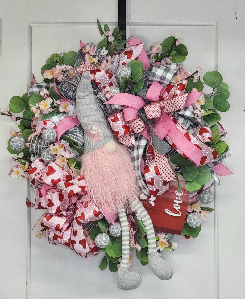 May include: A Valentine's Day wreath with a gnome figure. The wreath is decorated with pink and red heart-patterned ribbons, pink bows, and artificial foliage. A small red sign displays the word "Love".