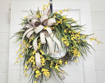 May include: A spring wreath with a burlap bow and yellow flowers. The wreath is made of green branches and is hanging on a white door.