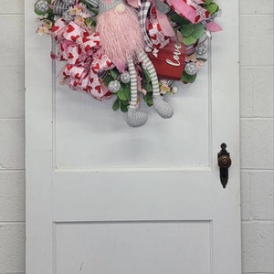 May include: A Valentine's Day wreath with a gnome figure. The wreath features pink and red ribbons, heart-shaped accents, and greenery. The gnome has a grey hat, pink beard, and striped legs. A small red box with the word "love" is included.