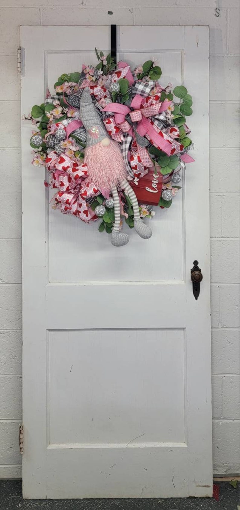 May include: A Valentine's Day wreath with a gnome figure. The wreath features pink and red ribbons, heart-shaped accents, and greenery. The gnome has a grey hat, pink beard, and striped legs. A small red box with the word "love" is included.