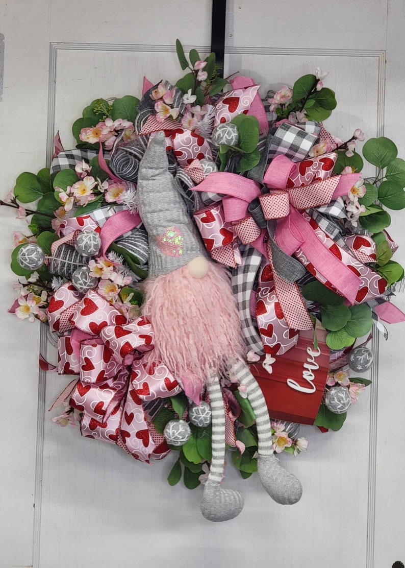 May include: A Valentine's Day wreath with a gnome figure. The wreath is decorated with pink and red heart-patterned ribbons, grey and white checked ribbons, and faux greenery. A red sign displays the word "Love".