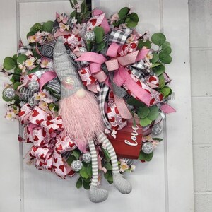 May include: A Valentine's Day wreath with a gnome figure. The wreath features pink and red ribbons with heart patterns, green foliage, and small white flowers. A red box with the word "love" is also included.