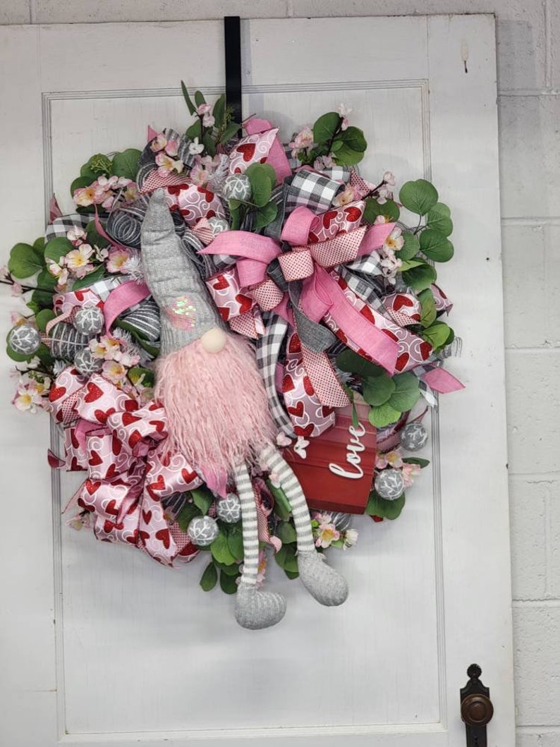 May include: A Valentine's Day wreath with a gnome figure. The wreath features pink and red ribbons with heart patterns, green foliage, and small white flowers. A red box with the word "love" is also included.