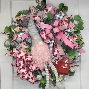 May include: A Valentine's Day wreath with a gnome figure. The wreath is adorned with pink and red heart-patterned ribbons, grey and white checked ribbons, and artificial flowers. A small red sign displays the word "love".