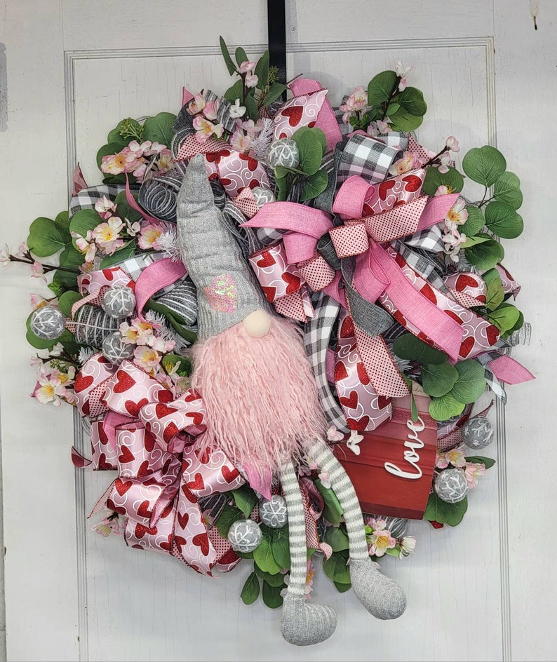 May include: A Valentine's Day wreath with a gnome figure. The wreath is adorned with pink and red heart-patterned ribbons, grey and white checked ribbons, and artificial flowers. A small red sign displays the word "love".