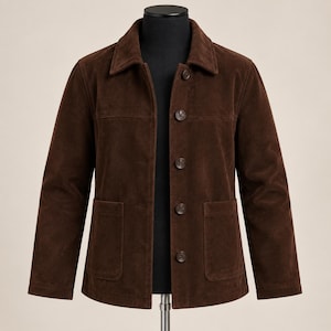 May include: A dark brown suede jacket featuring a classic collar, long sleeves, and a front button closure with five matching brown buttons. The jacket includes two large patch pockets on the lower front and horizontal seam detailing across the chest.