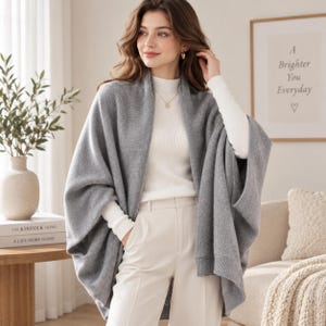 May include: A grey knit open-front poncho worn over a white long-sleeve ribbed mock-neck top and white trousers. The poncho features a soft texture, wide sleeves, and a draped silhouette that falls loosely over the shoulders and torso.
