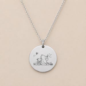 Op de afbeelding: Een zilverkleurige ronde hanger aan een ketting met een fijne zwarte lijntekening van een beer en een klein dier die samen in een veld zitten, met een vliegende bij en een paardenbloem, hangend aan een dunne zilveren ketting.
