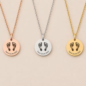 Op de afbeelding: Drie ronde kettinghangers in roségoud, zilver en goud. Elke hanger is voorzien van een zwarte gravure van twee babyvoetjes, de naam Sara en de datum 02.04.2025, hangend aan bijpassende, verfijnde metalen kettingen.