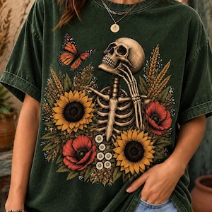 May include: A dark green mineral-washed t-shirt featuring a graphic print of a human skeleton surrounded by yellow sunflowers, red poppies, wheat stalks, and small white daisies, with an orange and black monarch butterfly near the skull.