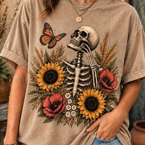 May include: A tan short-sleeve t-shirt featuring a graphic print of a human skeleton surrounded by yellow sunflowers, red poppies, and wheat stalks. An orange and black monarch butterfly is positioned near the skull, which is resting on a hand.
