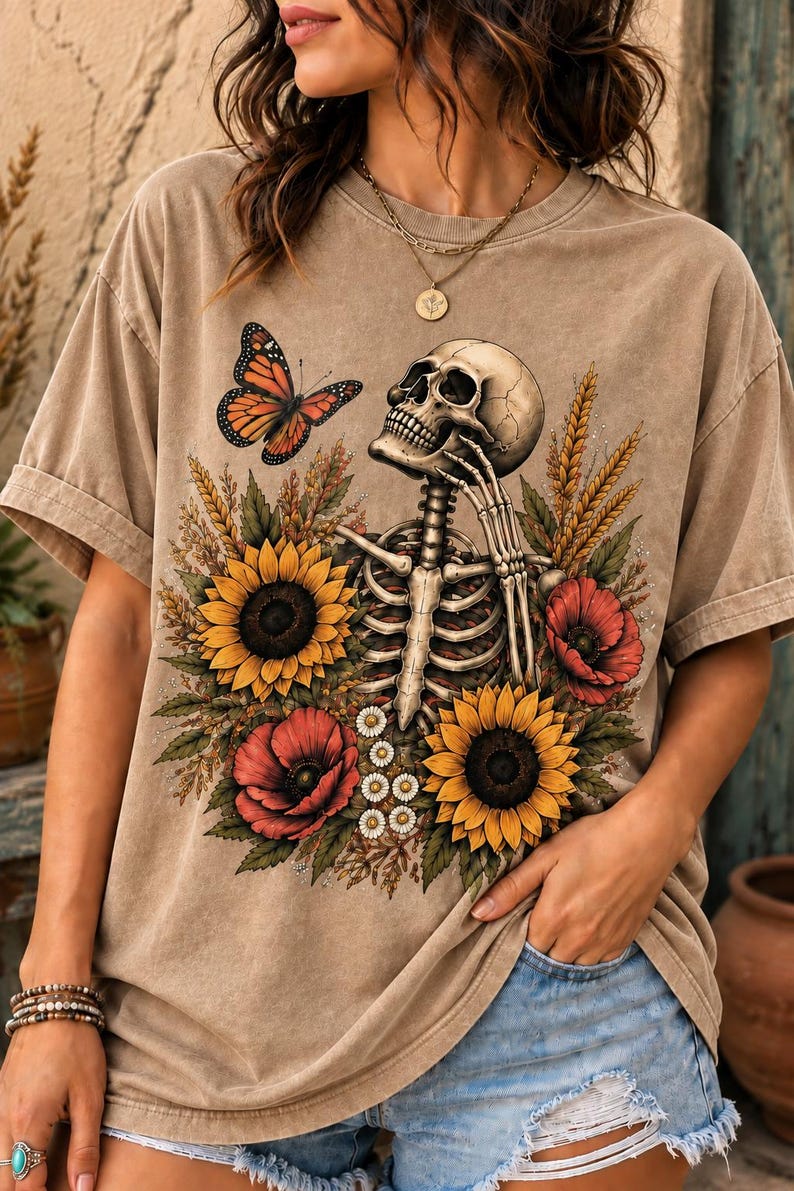 May include: A tan short-sleeve t-shirt featuring a graphic print of a human skeleton surrounded by yellow sunflowers, red poppies, and wheat stalks. An orange and black monarch butterfly is positioned near the skull, which is resting on a hand.