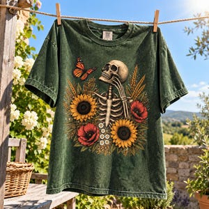 May include: A dark green short-sleeve t-shirt featuring a graphic print of a human skeleton surrounded by yellow sunflowers, red poppies, and a monarch butterfly. The skeleton is positioned with one hand near its chin, set against a floral background.