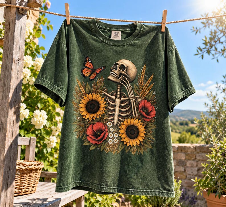 May include: A dark green short-sleeve t-shirt featuring a graphic print of a human skeleton surrounded by yellow sunflowers, red poppies, and a monarch butterfly. The skeleton is positioned with one hand near its chin, set against a floral background.