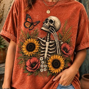 May include: A rust-colored mineral-washed t-shirt featuring a graphic print of a human skeleton surrounded by yellow sunflowers, red poppies, and green foliage. A monarch butterfly is positioned near the skull, which rests on a hand.