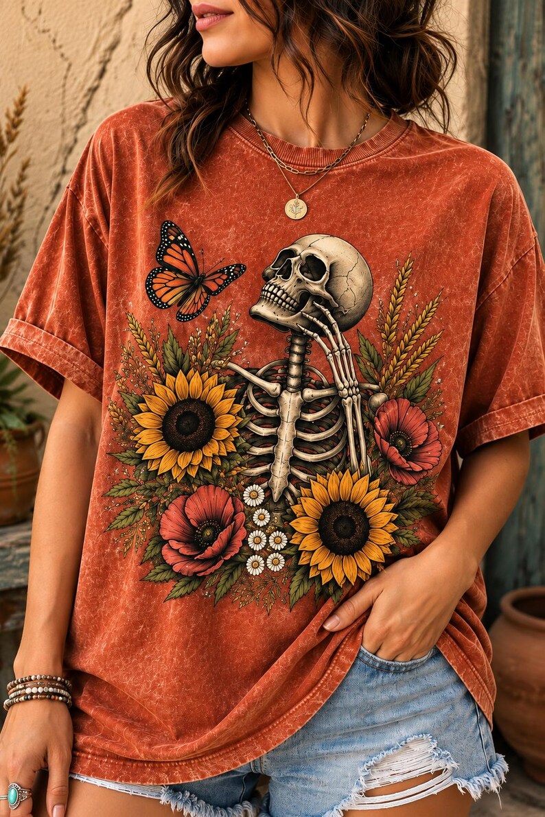 May include: A rust-colored mineral-washed t-shirt featuring a graphic print of a human skeleton surrounded by yellow sunflowers, red poppies, and green foliage. A monarch butterfly is positioned near the skull, which rests on a hand.