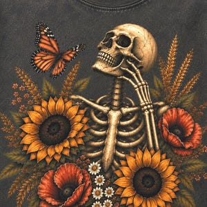 May include: A charcoal gray t-shirt featuring a graphic print of a human skeleton resting its chin on its hand. The skeleton is surrounded by vibrant yellow sunflowers, red poppies, small white daisies, and a single orange monarch butterfly.