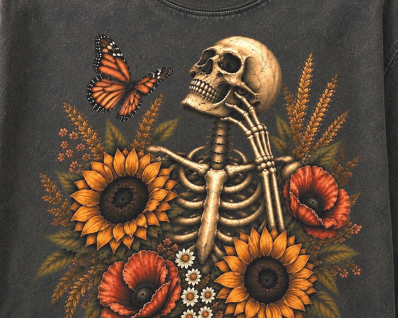 May include: A charcoal gray t-shirt featuring a graphic print of a human skeleton resting its chin on its hand. The skeleton is surrounded by vibrant yellow sunflowers, red poppies, small white daisies, and a single orange monarch butterfly.