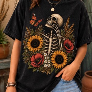 May include: A black short-sleeve t-shirt featuring a graphic print of a human skeleton surrounded by yellow sunflowers, red poppies, white daisies, wheat stalks, and an orange butterfly. The shirt has a crew neckline and a relaxed, loose fit.