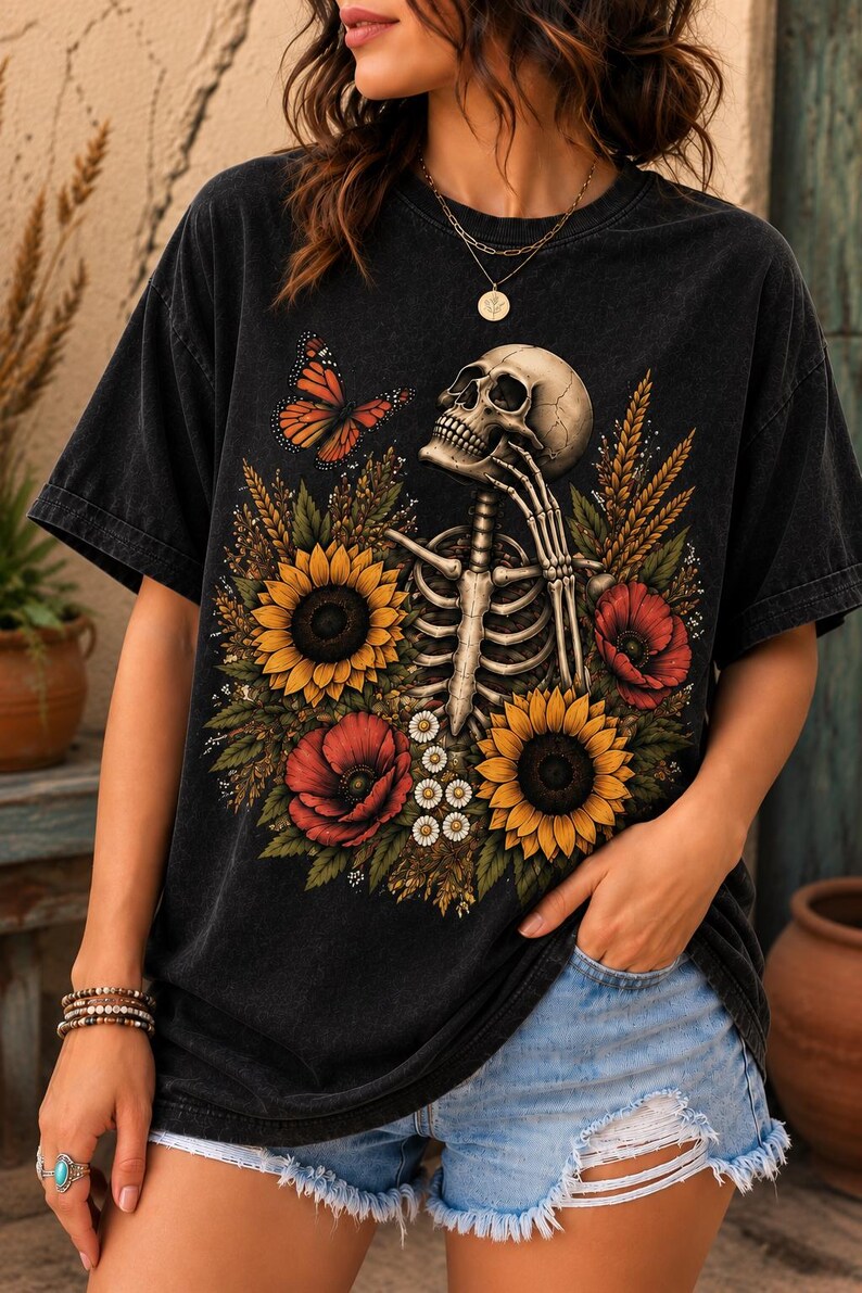 May include: A black short-sleeve t-shirt featuring a graphic print of a human skeleton surrounded by yellow sunflowers, red poppies, white daisies, wheat stalks, and an orange butterfly. The shirt has a crew neckline and a relaxed, loose fit.