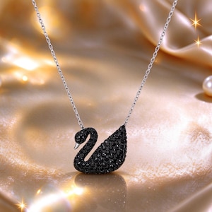May include: A silver-toned chain necklace featuring a swan-shaped pendant. The swan is encrusted with numerous small, dark black crystals and has a smooth, polished silver-coloured beak. The necklace hangs from a delicate, thin metal link chain.