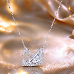 May include: A silver-toned necklace featuring a swan-shaped pendant. The swan is encrusted with numerous small, clear, sparkling stones and features a larger, marquise-cut clear stone at the centre of its wing. The pendant hangs from a delicate chain.