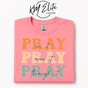 May include: A coral pink crew neck t-shirt featuring the text PRAY on it, PRAY over it, PRAY through it in stacked, wavy, retro-style lettering. The words are printed in orange, light peach, and sage green, with cursive black script text.