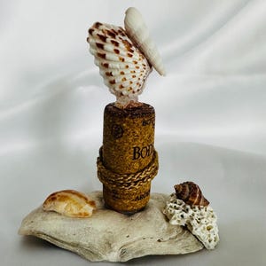 May include: A decorative cork stopper featuring a brown and white patterned seashell on top, wrapped with a thin gold cord. The cork is mounted on a large, flat, light-colored seashell base, accompanied by a small brown shell and white coral.