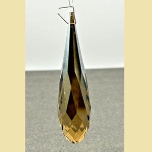 May include: A faceted, teardrop-shaped crystal pendant. The crystal is a gradient of brown and gold hues, suspended by a thin wire. The pendant's geometric cuts reflect light, creating a sparkling effect. Ideal for jewelry or decorative purposes.