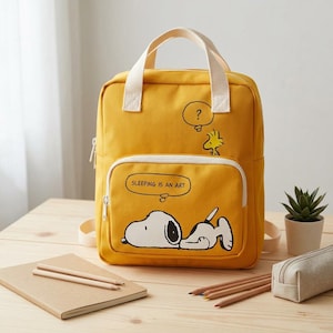 May include: A mustard yellow backpack featuring a graphic of Snoopy lying down with the text SLEEPING IS AN ART in a speech bubble and Woodstock with a question mark. The bag has two top handles, a front zippered pocket, and off-white straps.