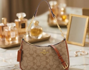 May include: A tan and brown monogram print handbag featuring a gold-tone chain handle, a detachable leather strap, and a gold Coach logo on the front. The bag includes a hanging leather tag and gold-tone hardware accents throughout the design.