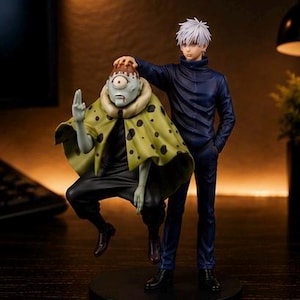 May include: A collectible figure set featuring a character with white hair in a dark blue high-collar uniform standing next to a smaller, pale-skinned creature with one eye, wearing a yellow cape with black spots and a dark outfit on a base.