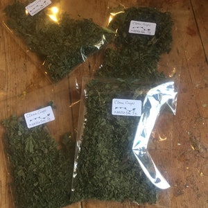 Könnte beinhalten: Vier durchsichtige Plastiktüten gefüllt mit getrockneten grünen Brennnesselblättern. Jede Tüte hat ein weißes Etikett mit dem Text "Clean Crops" und "Nettle tea 5g".