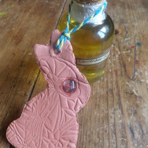Puede incluir: Una pequeña botella de vidrio transparente con un tapón de corcho y una cuerda azul y amarilla atada alrededor del cuello. La botella está llena de un líquido amarillo claro y tiene una etiqueta que dice "Lavender Oil". La botella está sentada sobre una superficie de madera junto a un adorno de terracota en forma de conejo.