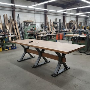 Puede incluir: Una mesa rectangular larga de madera con una superficie lisa de color marrón claro y una base robusta construida con patas de metal gris oscuro en forma de X, conectadas por una viga de soporte horizontal de madera.