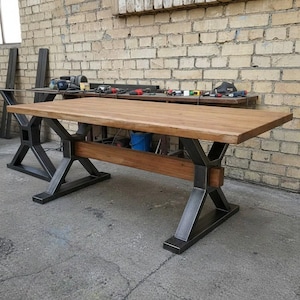 May include: A rectangular dining table featuring a thick, natural-toned wooden tabletop and a sturdy base constructed from dark gray metal. The base consists of two X-shaped supports connected by a horizontal wooden beam running between the legs.