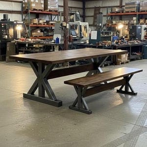 Puede incluir: Una mesa de comedor rectangular y un banco a juego, ambos con tableros de madera marrón oscuro y bases metálicas robustas en forma de X. El conjunto de muebles está construido con tablones de madera gruesos y soportes de acero.