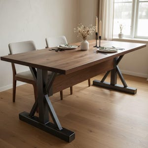 Puede incluir: Una mesa de comedor rectangular con tablero de madera marrón oscuro y base de metal negro en forma de X. La mesa está puesta con dos platos blancos, cubiertos negros, dos candelabros negros con velas color crema y un jarrón con flores secas.
