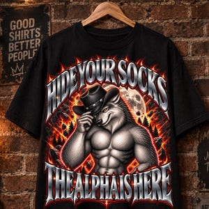 Pode incluir: Uma t-shirt preta com um gráfico de um lobo musculado a usar um chapéu de cowboy. O texto HIDE YOUR SOCKS está impresso acima do lobo e THE ALPHA IS HERE abaixo. O design inclui um fundo laranja e vermelho ardente.