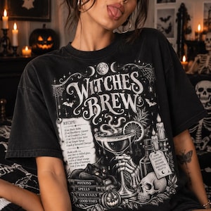 Op de afbeelding: Een zwart t-shirt met een witte grafische print getiteld Witches Brew. Het ontwerp bevat een cocktailrecept, een skeletthand die een glas vasthoudt, een zwarte kat op boeken, een schedel, kaarsen en de tekst Potions, Spells, Cocktails, Good Times.