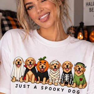 Könnte beinhalten: Ein weißes T-Shirt mit einer Grafik von sechs Golden Retrievern in verschiedenen Halloween-Kostümen, darunter ein Geist, ein Vampir, ein Kürbis, eine Mumie, ein Skelett und Frankenstein, mit dem darunter gedruckten Text "JUST A SPOOKY DOG".