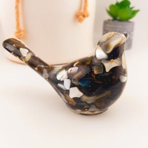 May include: A bird figurine crafted from dark resin embedded with iridescent shell fragments and small gold flecks. The smooth, glossy surface features a mosaic-like pattern of black, brown, and white tones, capturing the shape of a small songbird.