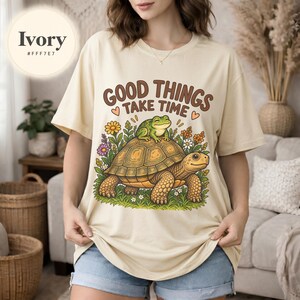 Pode incluir: Uma t-shirt cor de marfim com uma estampa gráfica de uma tartaruga castanha com um sapo verde no casco, rodeada por flores coloridas e relva verde. O texto "GOOD THINGS TAKE TIME" está impresso em castanho acima da ilustração.