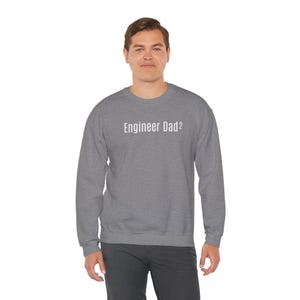 May include: A heather grey long-sleeve crewneck sweatshirt featuring the white text Engineer Dad² printed across the chest. The garment has a ribbed neckline, cuffs, and hem, providing a standard fit with long sleeves and a solid grey colour.