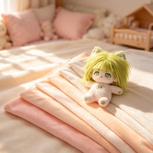 May include: A small doll with light green hair and cat ears sits on a stack of folded fabric pieces in shades of cream, peach, and light pink. The doll has large green eyes, a small mouth, and a green cat face embroidered on its chest.