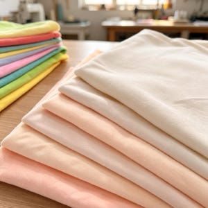 Op de afbeelding: Een stapel gevouwen stoffen in diverse pasteltinten waaronder crème, perzik en lichtroze, naast een aparte stapel met felgekleurde lagen stof in geel, groen, blauw en roze tinten.