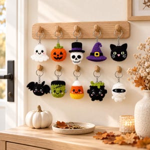 May include: A collection of ten crocheted Halloween-themed keychains hanging from a wooden wall rack. The figures include a white ghost, orange pumpkin, skull with a top hat, purple witch hat, black cat, black bat, green monster, candy corn, black cauldron, and a white mummy.